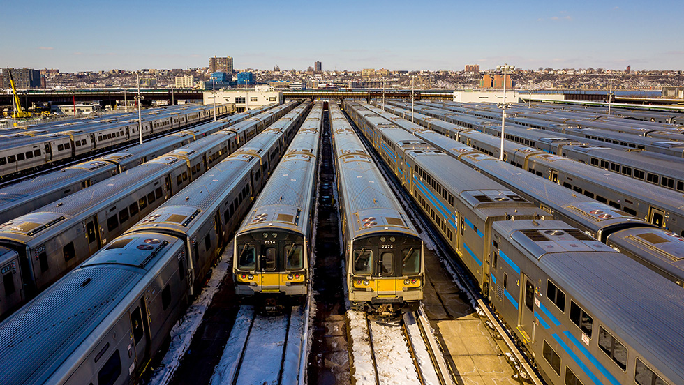 Rows of silver commuter train cars with blue stripes and yellow-fronted ends sit parked side by side in a rail yard on a clear day. Patches of snow cover the ground between the tracks, and a city skyline stands in the distance.