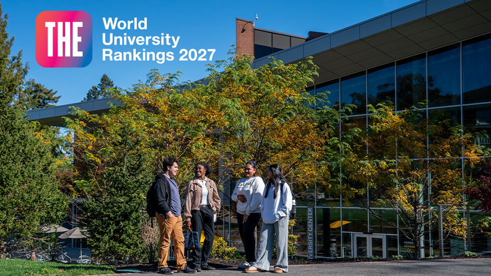 four students in casual attire standing in front of a modern university building surrounded by trees with fall foliage