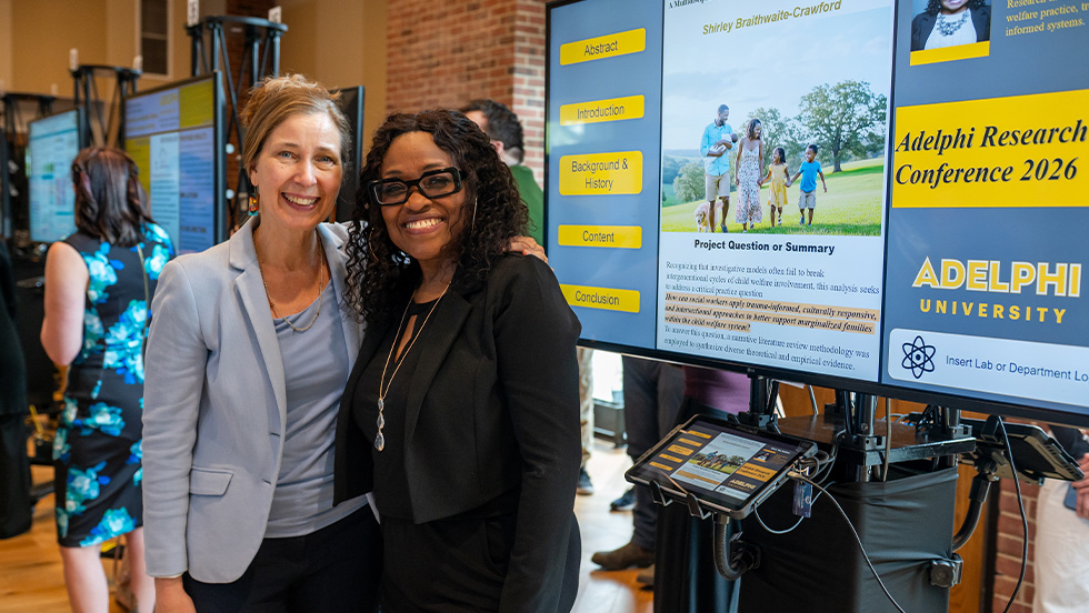 Two smiling women pose side by side with their arms around each other in front of a digital poster display at the Adelphi Research Conference 2026. One has light hair pulled back and wears a gray blazer, and the other has dark curly hair, glasses, and a black blazer. The screen behind them shows a research poster with a photo of a family walking outdoors, and other poster displays are visible in the background.