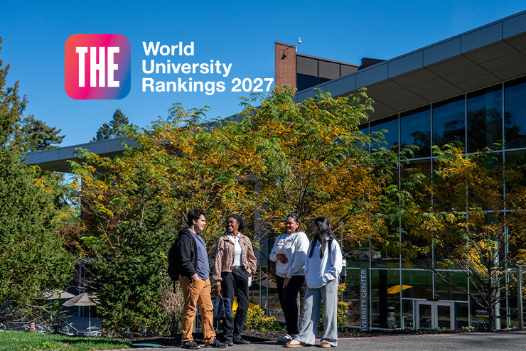 Four students in casual attire standing in front of a modern university building surrounded by trees with fall foliage. Badge for 2027 Times Higher Education (THE) World University Rankings.