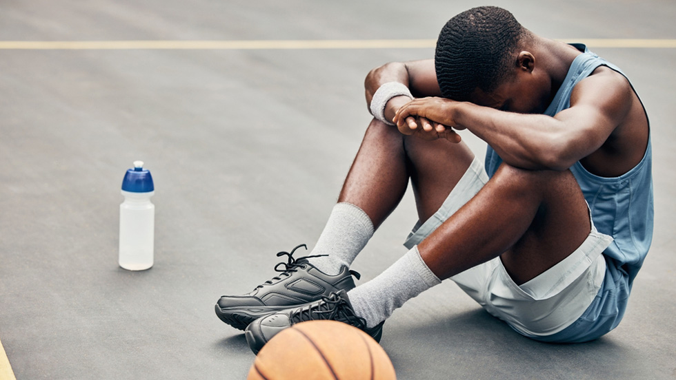 A young male basketball player sits on an outdoor court with his head down, resting on his arms folded over his knees, next to a water bottle and basketball.