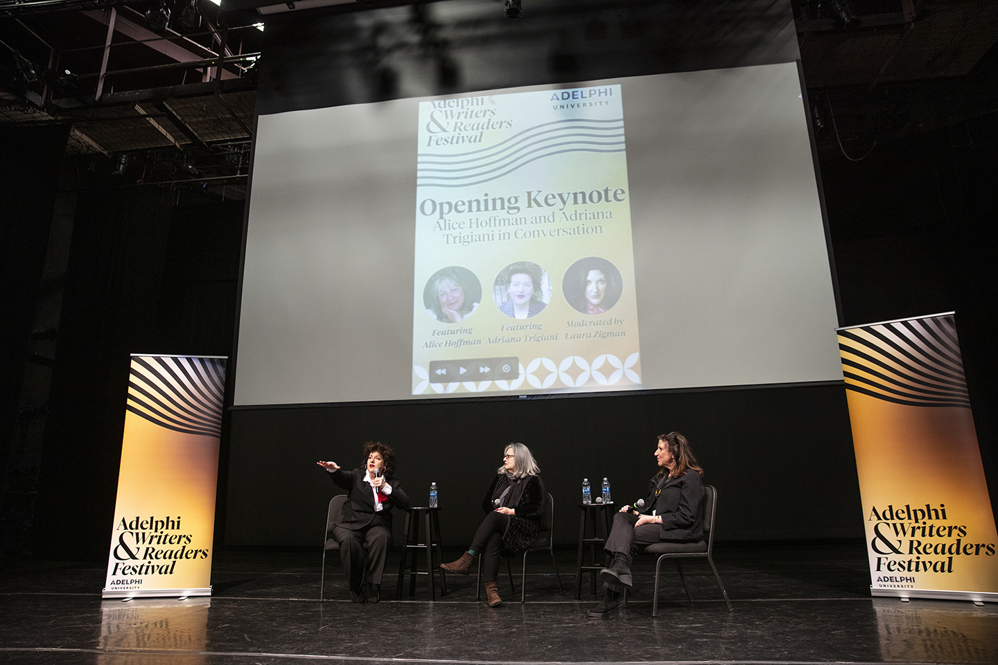 Authors and moderator seated on stage. There are banners on each side of them that read “Adelphi Writers & Readers Festival” and a screen above them that says “Opening Keynote: Alice Hoffman and Adriana Trigiani in Conversation.” The screen also has their photos, along with a photo of the moderator.