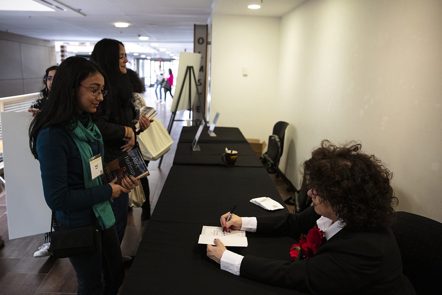 The writer seated at a long table, which is covered by a black tablecloth. She is signing a paperback copy of her book for an attendee, who is standing in front of the table. Other attendees are seen in the background.