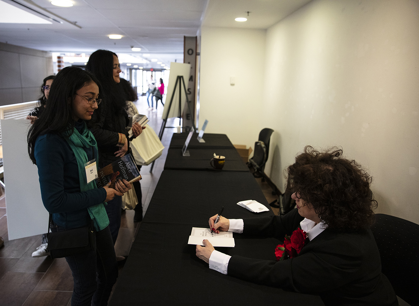 The writer seated at a long table, which is covered by a black tablecloth. She is signing a paperback copy of her book for an attendee, who is standing in front of the table. Other attendees are seen in the background.
