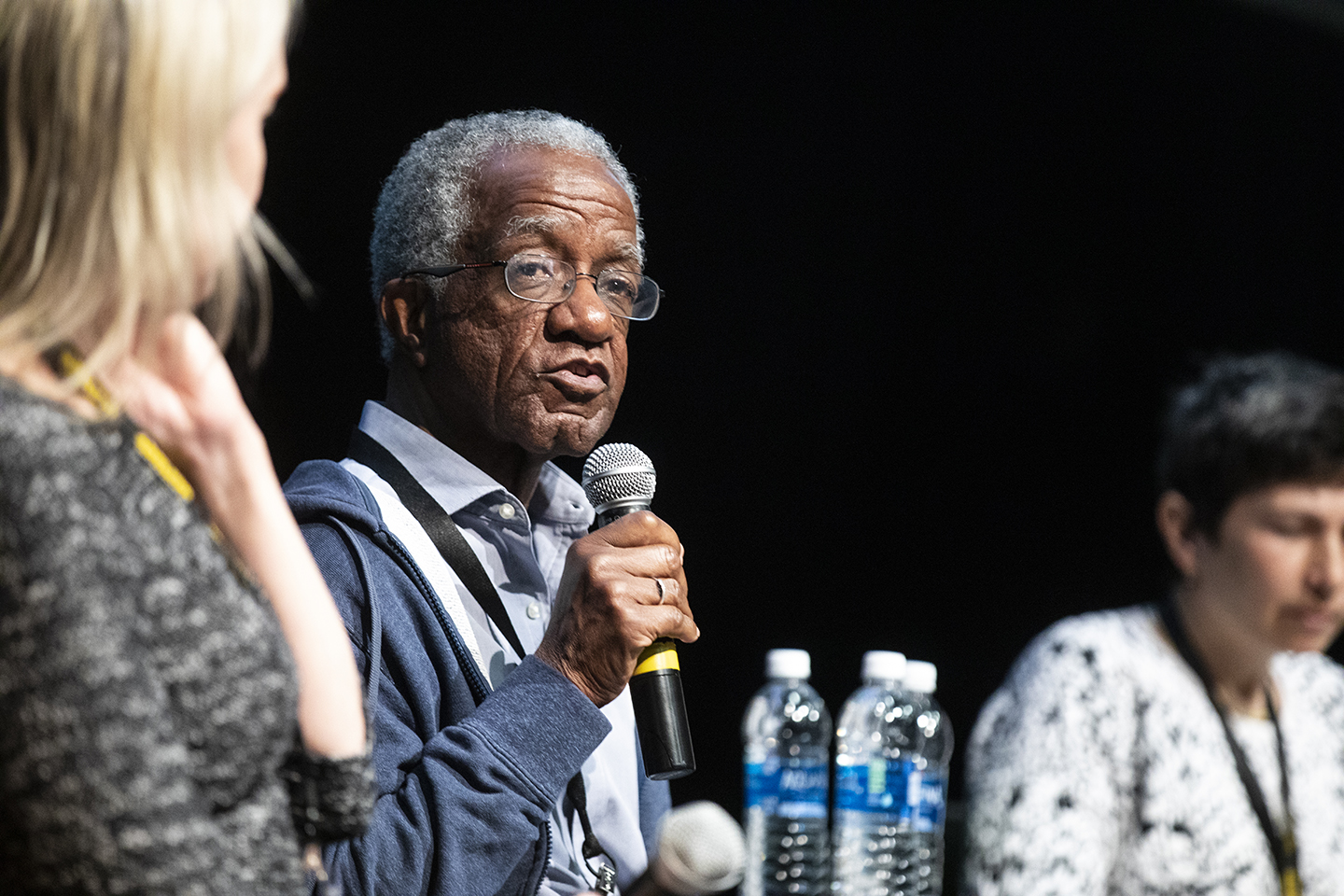 A close-up of the author, holding a microphone. Other panelists are shown but are out of focus.