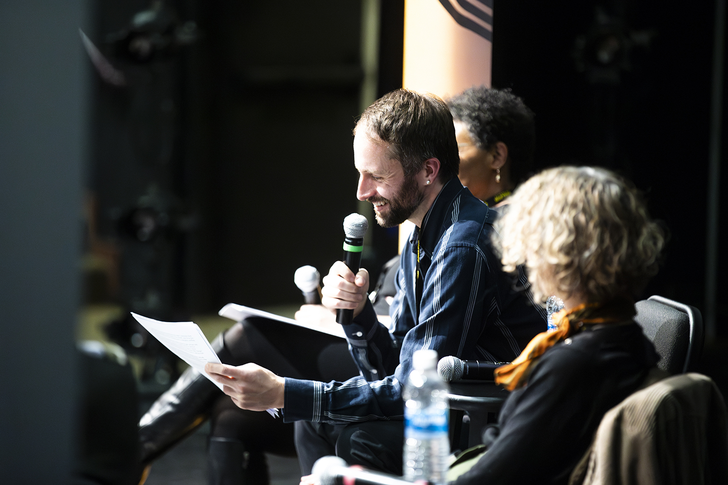 The poet on stage, seated and holding a microphone. He is flanked by two other panelists, who are out of focus.
